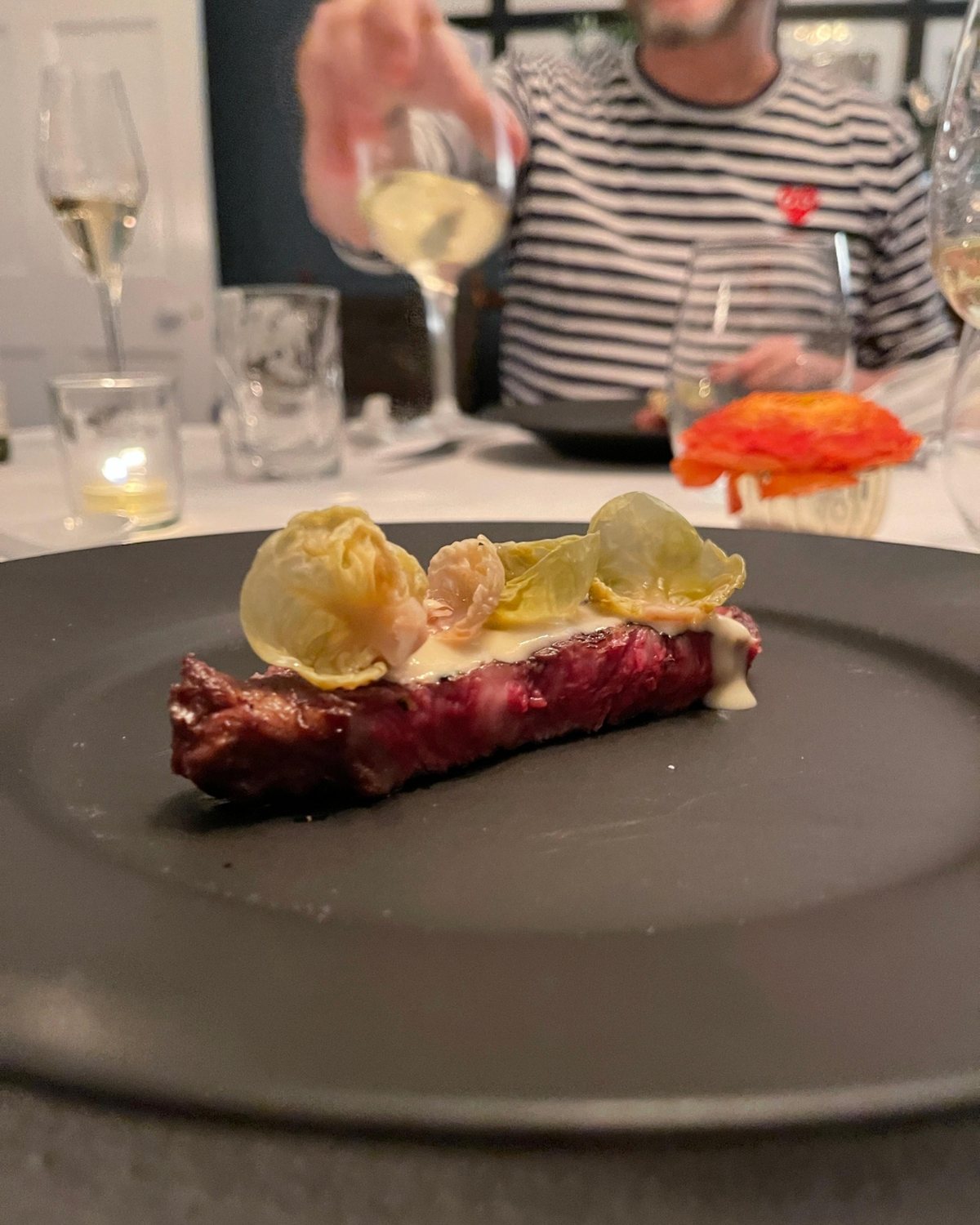 A seared beef course on a dark plate with charred brassica and cream sauce, candlelit at the supper table