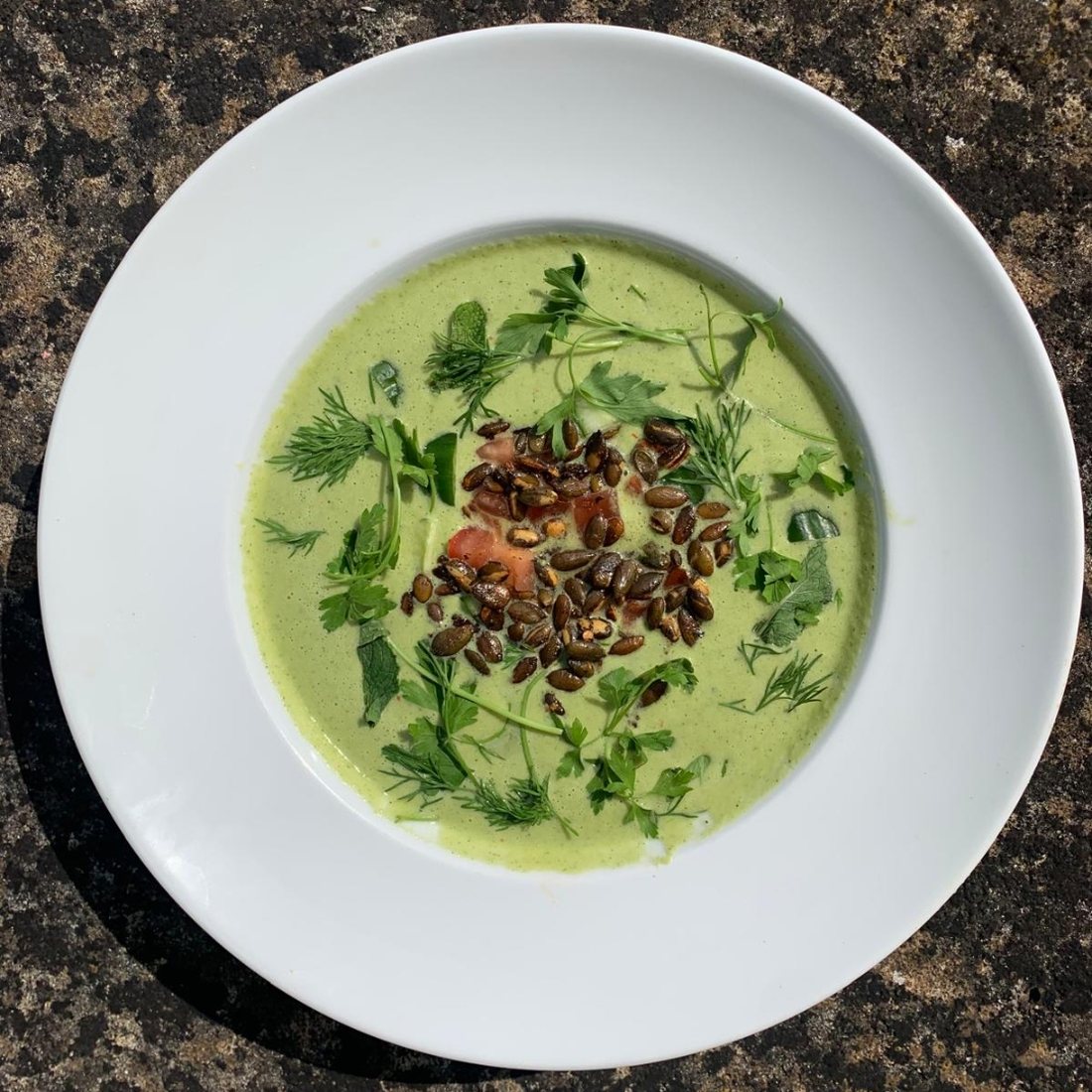 A chilled green herb soup in a wide white bowl, topped with toasted seeds, tomato and fresh herbs