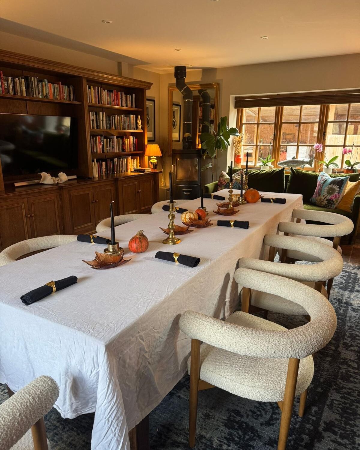 The long table set for supper with candles and bouclé chairs beside the wood-burner