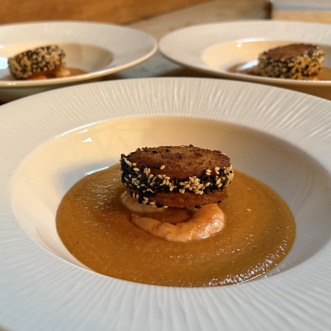 A smooth prawn and saffron bisque in a wide white bowl, topped with a sesame-crusted crouton and a whole prawn